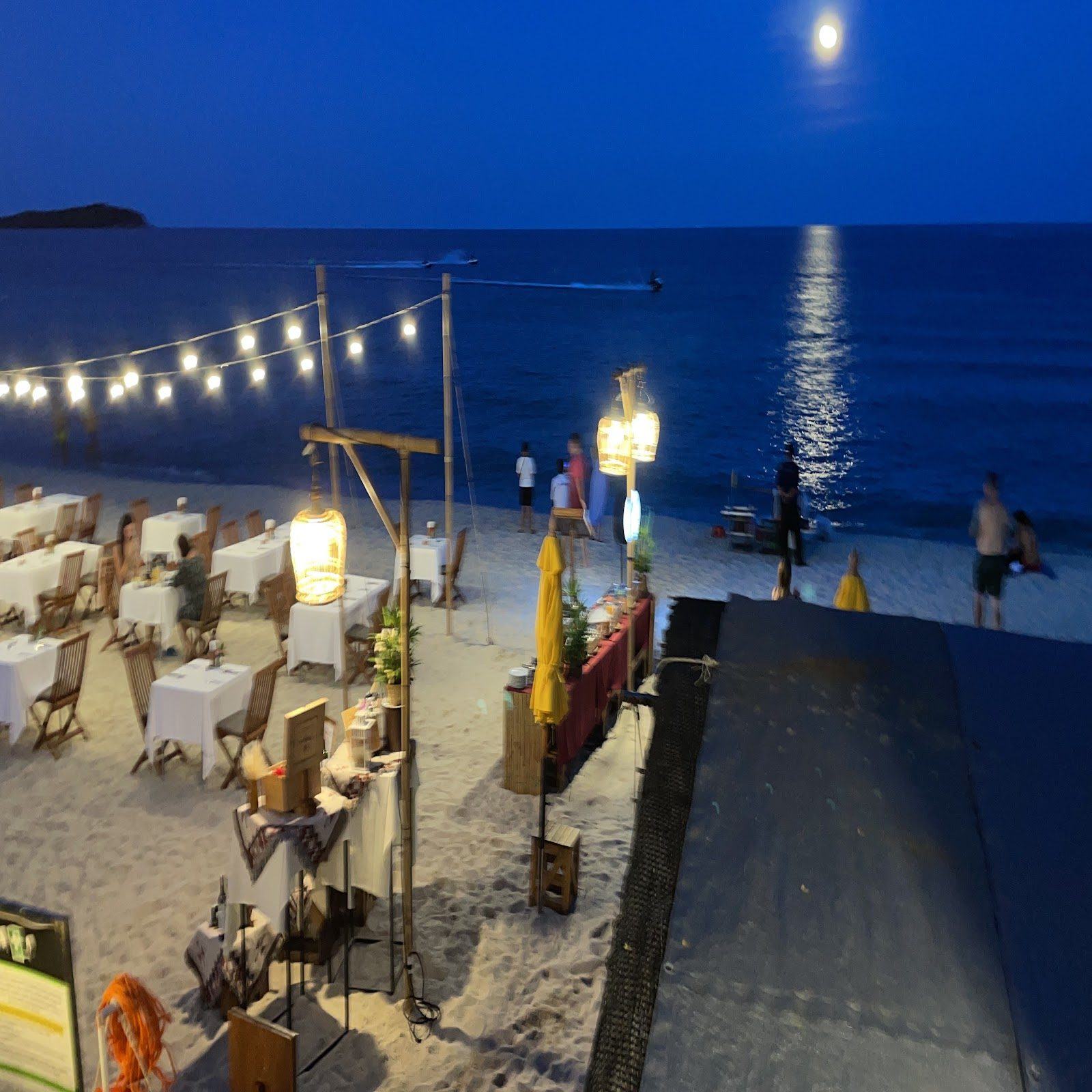 Beach restaurant tables under string lights and a full moon